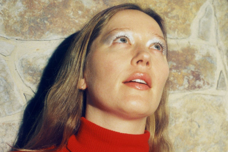 Close-up portrait of singer-songwriter Julia Jacklin in a red turtleneck sweater, wearing silver eyeshadow and looking upward against a stone wall.